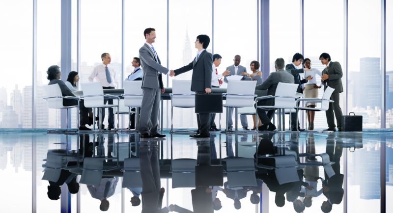 Imagen de un grupo de personas realizando una reunión de trabajo en una oficina vidriada que muestra en primer plano a dos hombres dándose la mano en señal de cerrar un trato