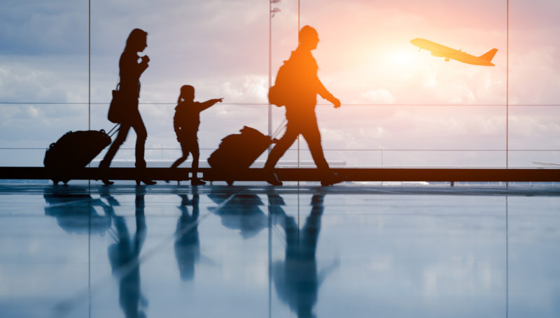 Imagen de una familia de tres personas caminanado por el aeropuerto con aviones despegando de fondo